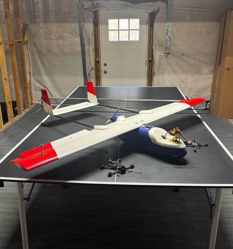 A large white and red fixed-wing aircraft on a table with two small racing quadcopters beside it.