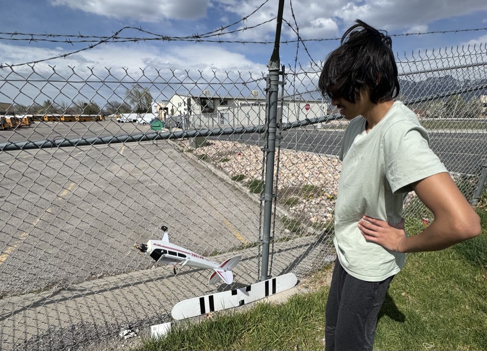 Looking down at a crashed white and red RC plane resting against a chain-link fence.