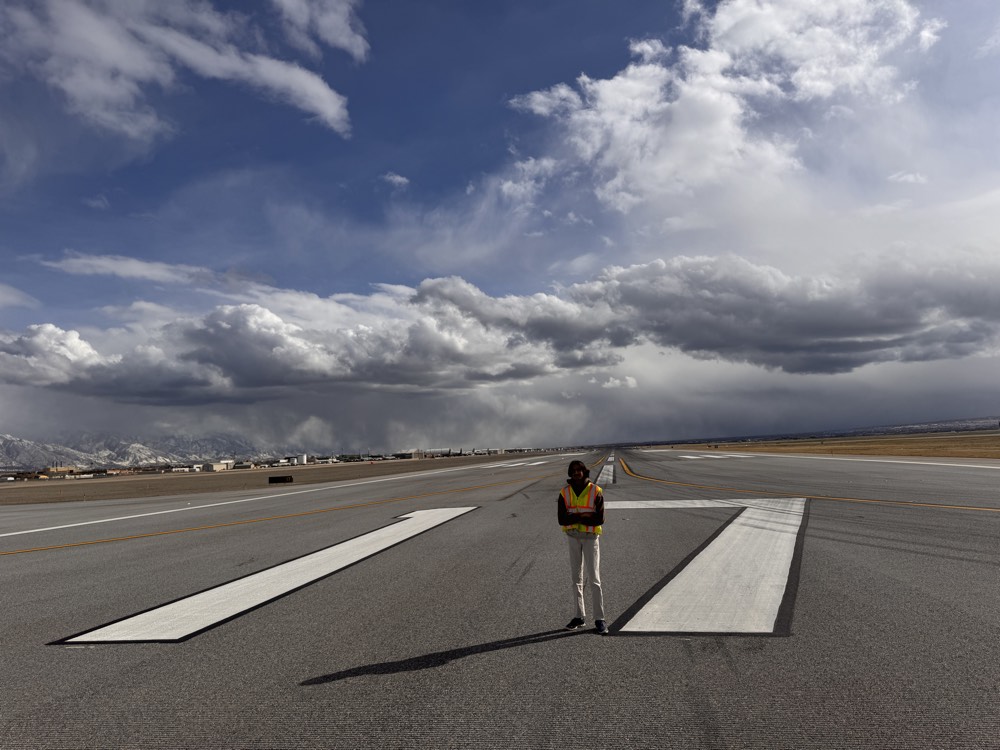 Standing in a high-visibility vest on an airport runway under heavy cloud, mountains in the distance.